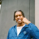 Nemzzz with braided hair wearing a blue jacket and pointing to his silver grills