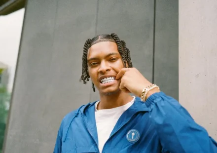 Nemzzz with braided hair wearing a blue jacket and pointing to his silver grills