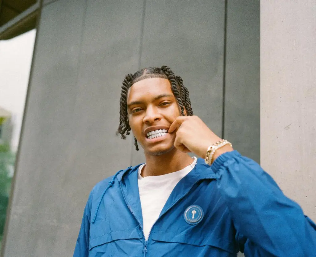 Nemzzz with braided hair wearing a blue jacket and pointing to his silver grills