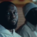 Headie One in a white hoodie looks directly forward, with Stormzy wearing a beanie in the dark background