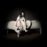 Billie Eilish sitting on a bed with white eyes and a grin, wearing all white in a dark room