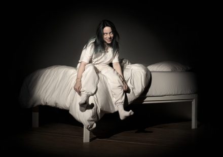 Billie Eilish sitting on a bed with white eyes and a grin, wearing all white in a dark room
