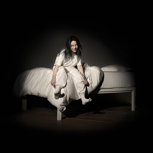 Billie Eilish sitting on a bed with white eyes and a grin, wearing all white in a dark room