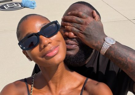 Rick Ross covering his eyes while posing for a selfie with a woman on an airport tarmac