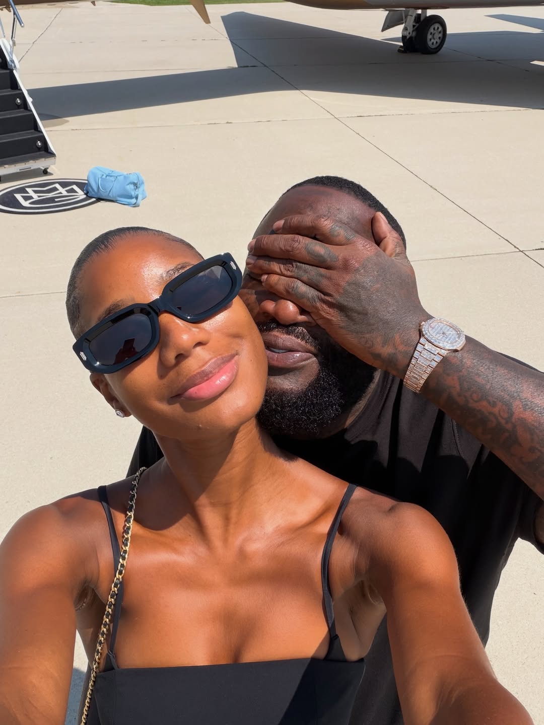 Rick Ross covering his eyes while posing for a selfie with a woman on an airport tarmac