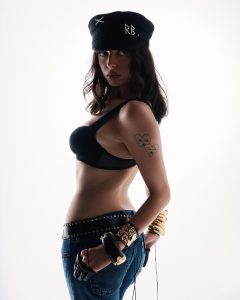 Woman in a black bra, jeans, and captain's hat with "RB" in pearls, looking over her shoulder