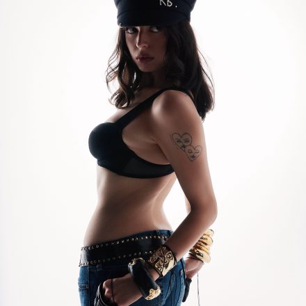 Woman in a black bra, jeans, and captain's hat with "RB" in pearls, looking over her shoulder
