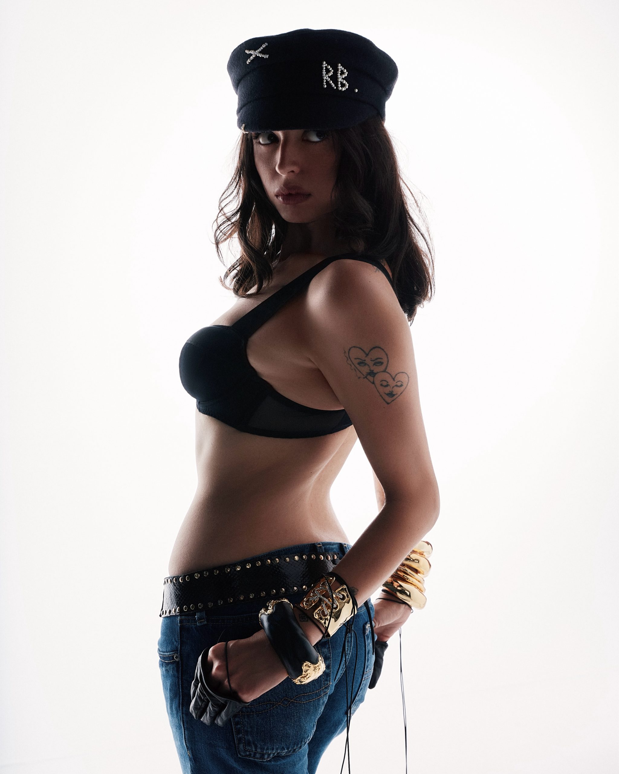 Woman in a black bra, jeans, and captain's hat with "RB" in pearls, looking over her shoulder