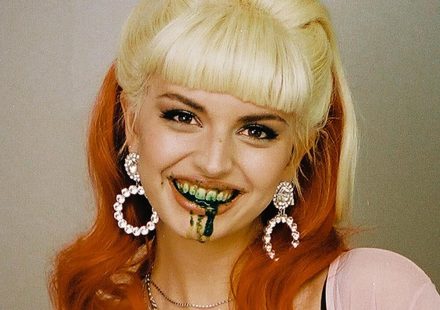 A person with blonde bangs and long orange hair smiles with dark green liquid dripping from their mouth
