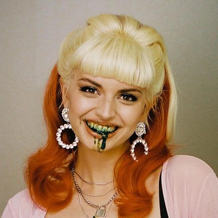 A person with blonde bangs and long orange hair smiles with dark green liquid dripping from their mouth