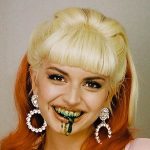 A person with blonde bangs and long orange hair smiles with dark green liquid dripping from their mouth