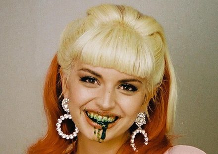 A person with blonde bangs and long orange hair smiles with dark green liquid dripping from their mouth