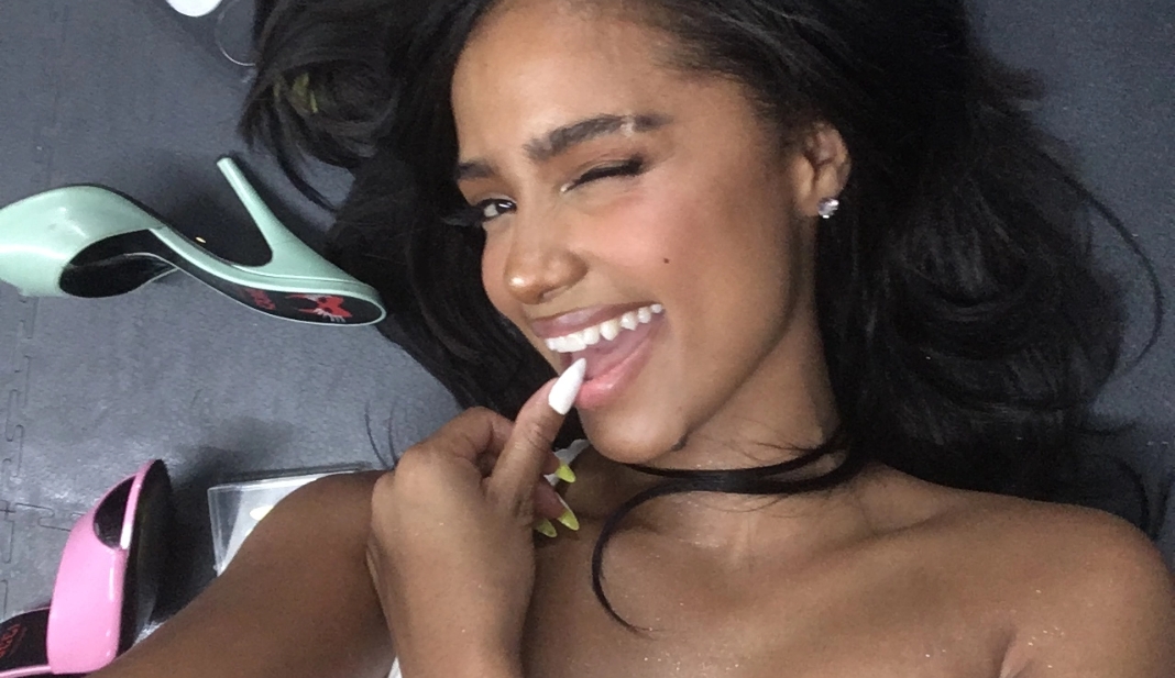 Tyla smiling with long dark hair winking whilst lying on the floor next to high-heeled shoes