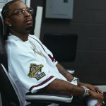 21 Savage sitting in a chair wearing a white 1995 World Series baseball jersey and round glasses
