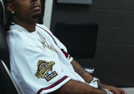 21 Savage sitting in a chair wearing a white 1995 World Series baseball jersey and round glasses
