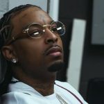 21 Savage sitting in a chair wearing a white 1995 World Series baseball jersey and round glasses