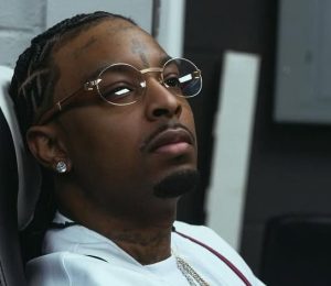 21 Savage sitting in a chair wearing a white 1995 World Series baseball jersey and round glasses