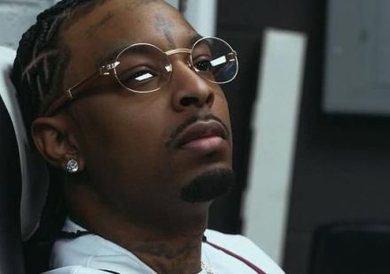 21 Savage sitting in a chair wearing a white 1995 World Series baseball jersey and round glasses