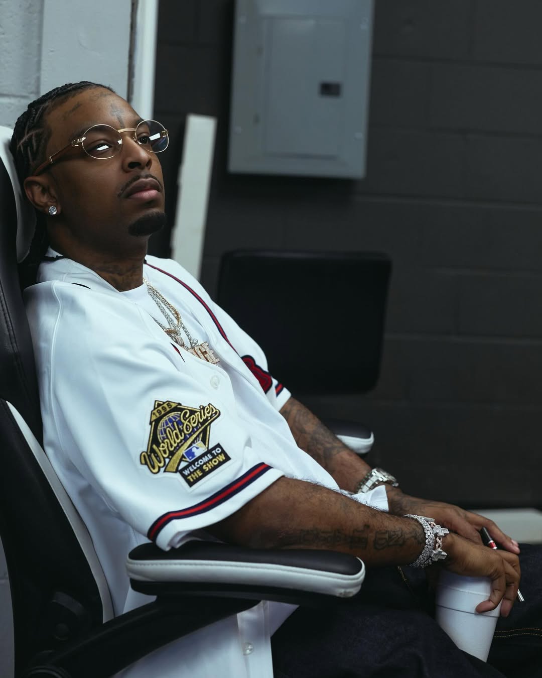 21 Savage sitting in a chair wearing a white 1995 World Series baseball jersey and round glasses