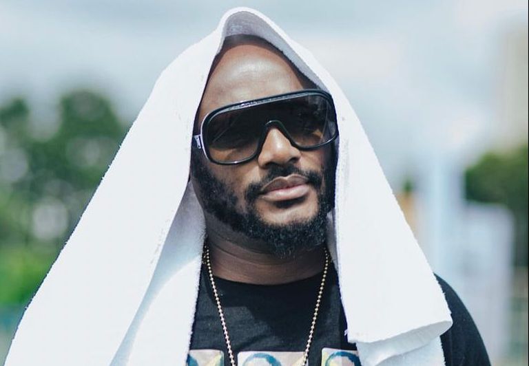 2Baba with a beard wearing large dark sunglasses, a gold chain, and a white towel draped over his head