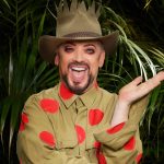 Boy George in a khaki shirt with red polka dots and a crown-topped hat, smiling in front of jungle foliage