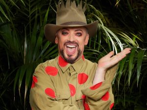 Boy George in a khaki shirt with red polka dots and a crown-topped hat, smiling in front of jungle foliage