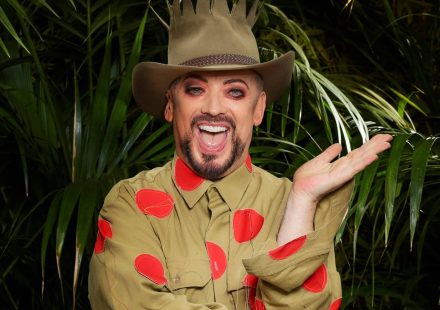 Boy George in a khaki shirt with red polka dots and a crown-topped hat, smiling in front of jungle foliage