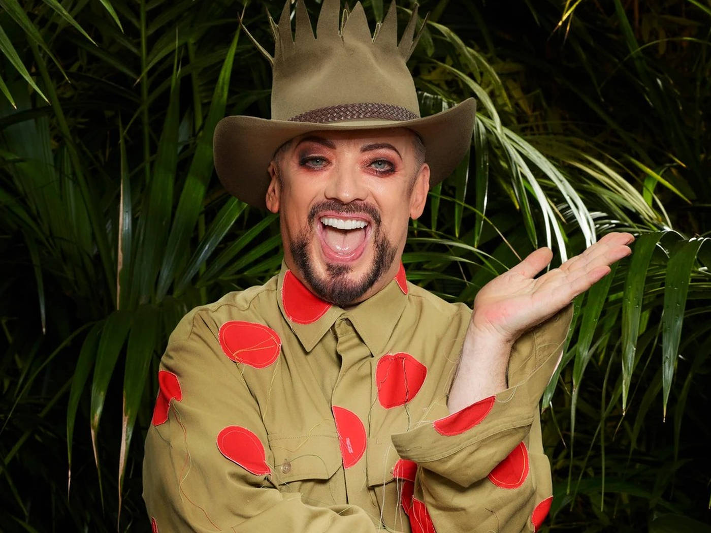 Boy George in a khaki shirt with red polka dots and a crown-topped hat, smiling in front of jungle foliage
