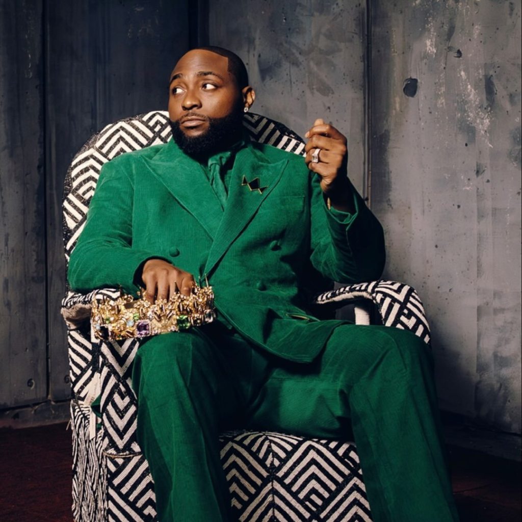 Davido sitting in a patterned chair wearing a green suit and holding a gold crown