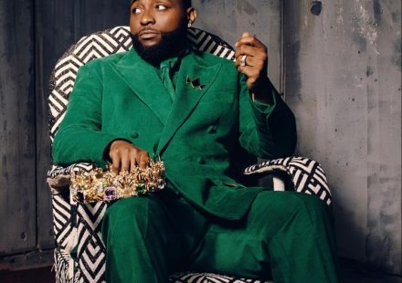 Davido sitting in a patterned chair wearing a green suit and holding a gold crown