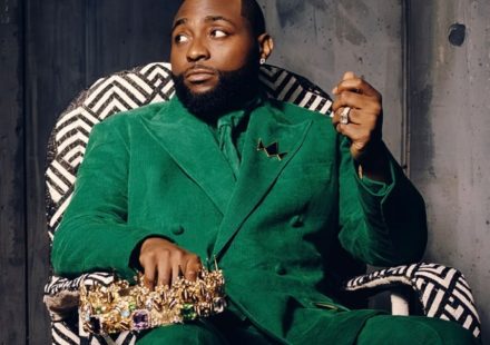 Davido sitting in a patterned chair wearing a green suit and holding a gold crown