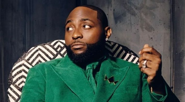 Davido sitting in a patterned chair wearing a green suit and holding a gold crown