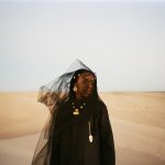 Dua Saleh wearing black clothing, gold jewelry, and a sheer black veil blowing in the wind amidst desert sand dunes