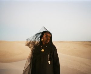 Dua Saleh wearing black clothing, gold jewelry, and a sheer black veil blowing in the wind amidst desert sand dunes