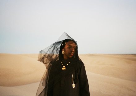 Dua Saleh wearing black clothing, gold jewelry, and a sheer black veil blowing in the wind amidst desert sand dunes