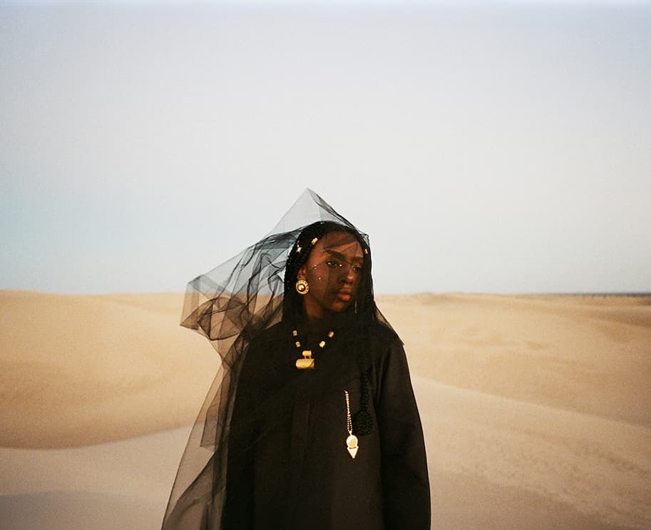 Dua Saleh wearing black clothing, gold jewelry, and a sheer black veil blowing in the wind amidst desert sand dunes