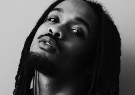 Black and white portrait of Jaïr Faria with long locks, a mustache, and goatee, wearing a necklace