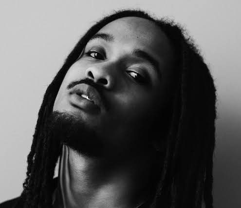Black and white portrait of Jaïr Faria with long locks, a mustache, and goatee, wearing a necklace