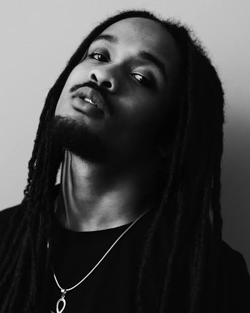Black and white portrait of Jaïr Faria with long locks, a mustache, and goatee, wearing a necklace
