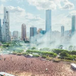 Aerial view of a massive crowd at a music festival in a city park with a backdrop of skyscrapers