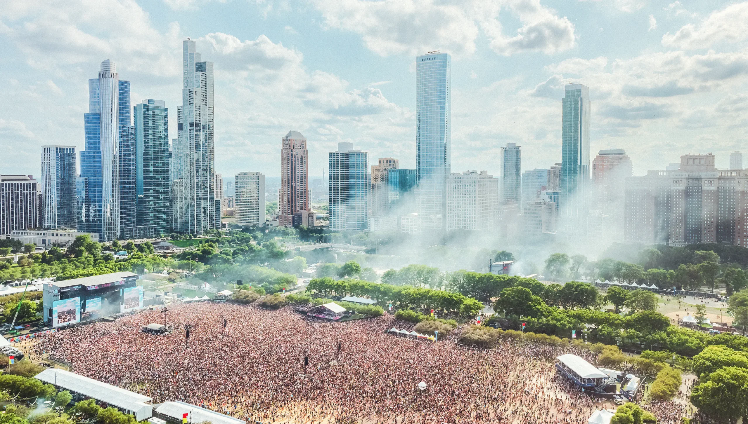 Aerial view of a massive crowd at a music festival in a city park with a backdrop of skyscrapers