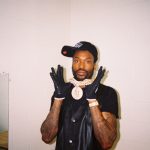 Meek Mill wearing a black cap, leather vest, and large diamond chain, posing with gloved hands up