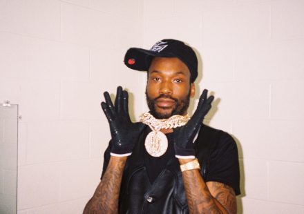 Meek Mill wearing a black cap, leather vest, and large diamond chain, posing with gloved hands up