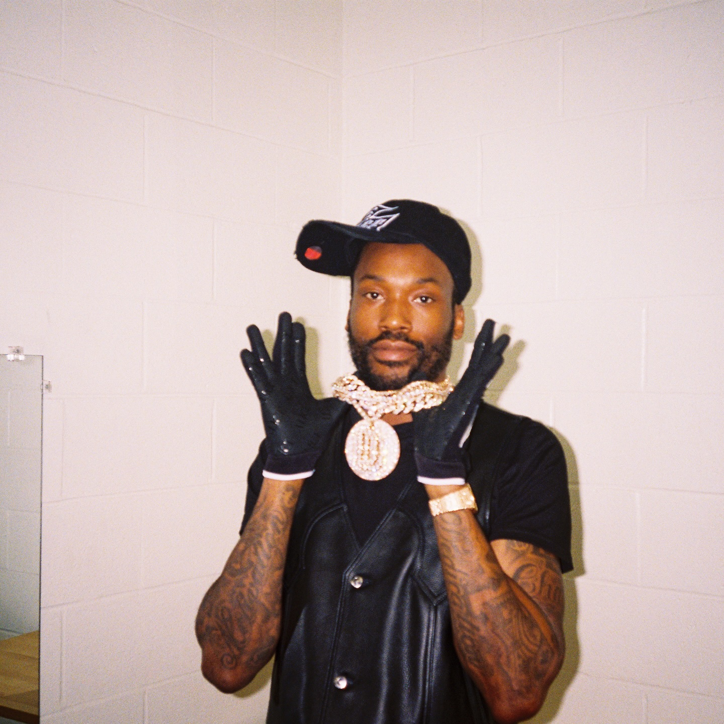Meek Mill wearing a black cap, leather vest, and large diamond chain, posing with gloved hands up