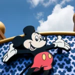 Mickey Mouse with arms outstretched in front of a blue wall patterned with Mickey silhouettes