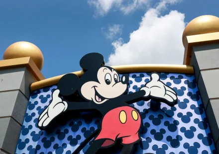 Mickey Mouse with arms outstretched in front of a blue wall patterned with Mickey silhouettes