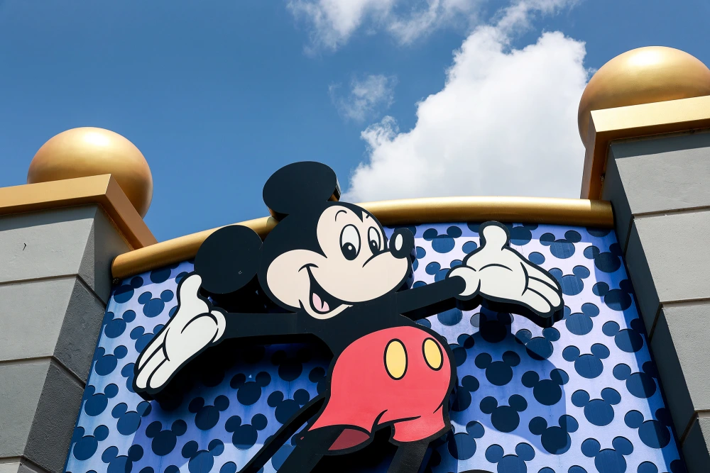 Mickey Mouse with arms outstretched in front of a blue wall patterned with Mickey silhouettes