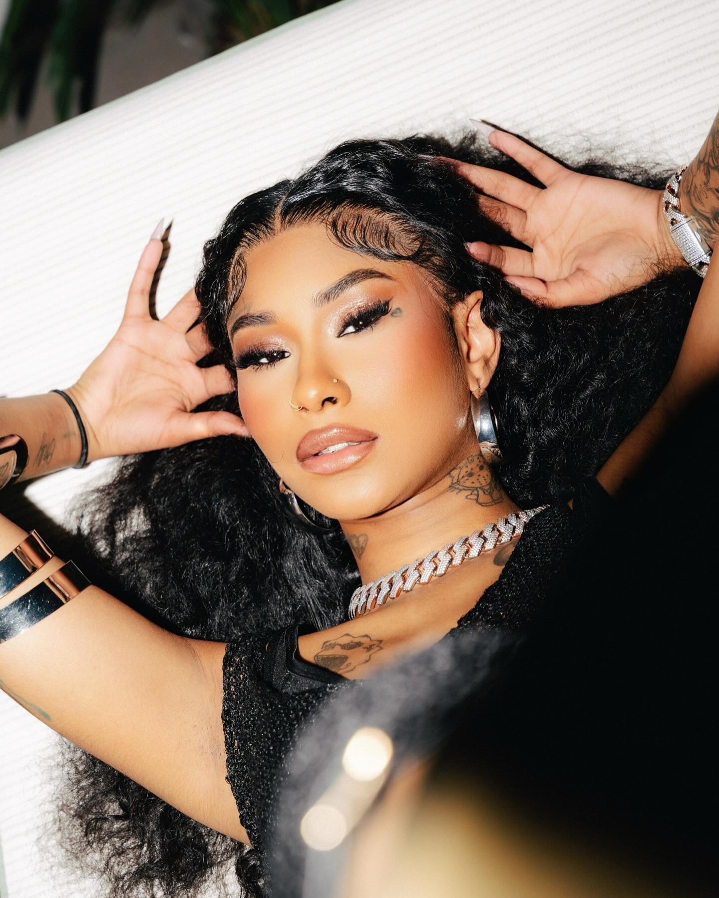 Ming Luanli with long curly hair and full makeup lying down, wearing a thick chain necklace and silver bracelets