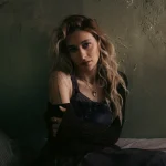 Paris Jackson with long wavy hair and shoulder tattoos sitting against a textured wall in dim lighting