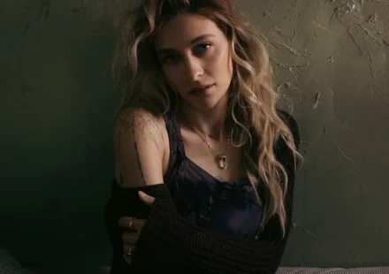 Paris Jackson with long wavy hair and shoulder tattoos sitting against a textured wall in dim lighting